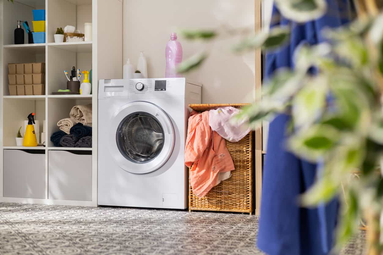 View of home laundry room, dresser with detergent, powder, towels, open washing machine with empty drum, next to wicker basket with colorful laundry items